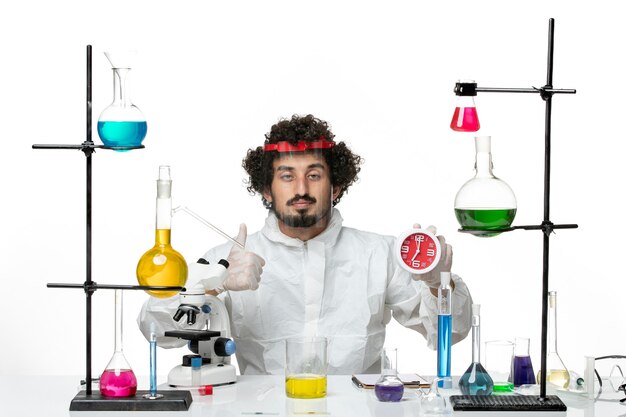 Vue de face jeune homme scientifique en costume spécial avec casque de protection tenant des horloges sur mur blanc science lab covid chimie male