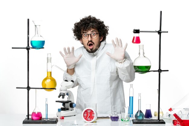 Vue de face jeune homme scientifique en costume spécial debout autour de la table avec des solutions sur un bureau blanc