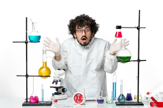 Vue de face jeune homme scientifique en costume spécial debout autour de la table avec des solutions sur le mur blanc