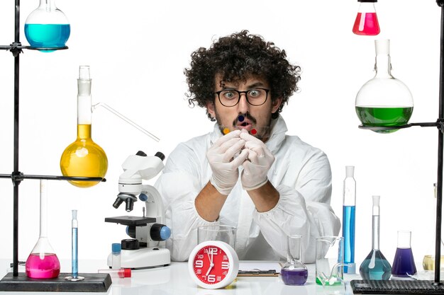Vue de face jeune homme scientifique en costume spécial travaillant autour de la table avec des solutions sur le mur blanc