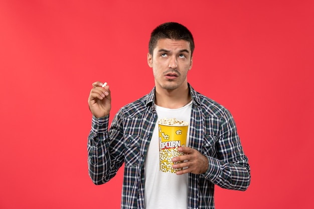 Vue de face jeune homme souriant et tenant le paquet de pop-corn et de la pensée sur le mur rouge clair hommes films cinéma cinéma film