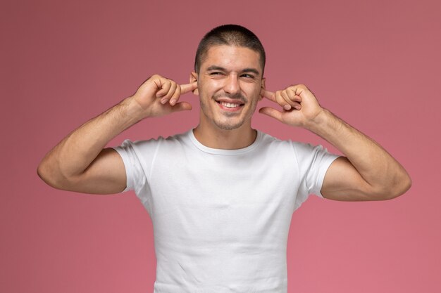 Vue de face jeune homme en t-shirt blanc collant ses oreilles avec sourire sur fond rose