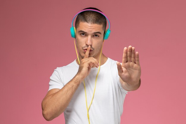 Vue de face jeune homme en t-shirt blanc à l'écoute de la musique showign silence signe sur bureau rose