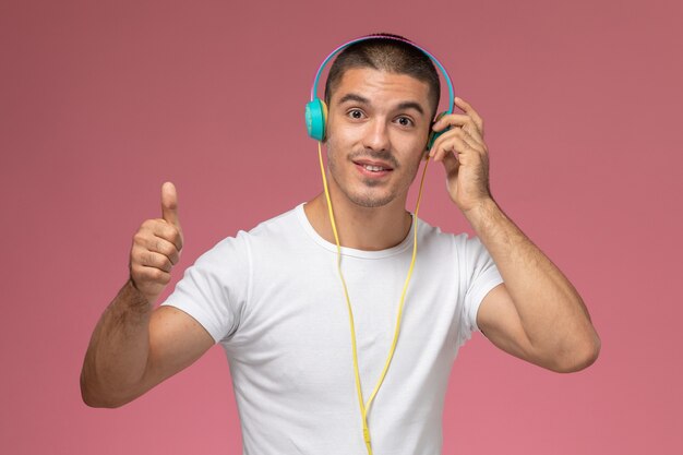 Vue de face jeune homme en t-shirt blanc, écouter de la musique via des écouteurs sur le bureau rose clair