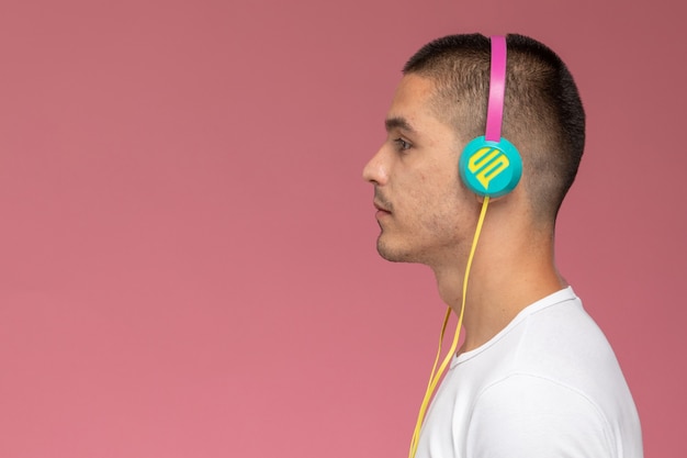 Photo gratuite vue de face jeune homme en t-shirt blanc, écouter de la musique via des écouteurs sur le fond rose clair