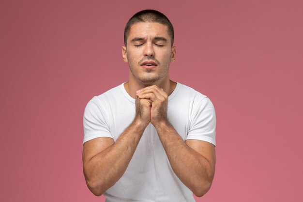 Vue de face jeune homme en t-shirt blanc en prière posant sur fond rose