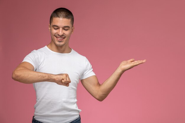 Vue de face jeune homme en t-shirt blanc regardant son poignet et souriant sur fond rose