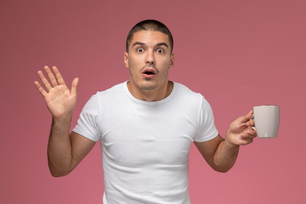 Vue de face jeune homme en t-shirt blanc tenant une tasse de café avec une expression surprise sur un bureau rose