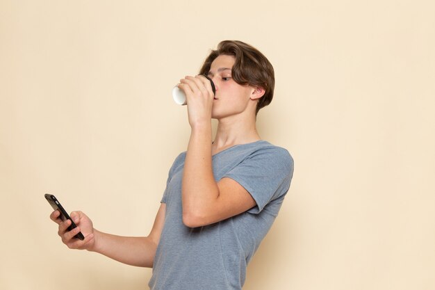 Une vue de face jeune homme en t-shirt gris, boire du café et à l'aide d'un téléphone