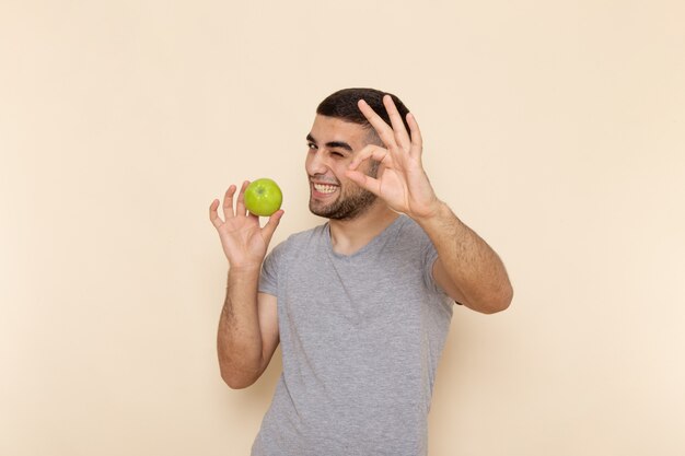 Vue de face jeune homme en t-shirt gris et jean bleu souriant et tenant une pomme verte sur beige