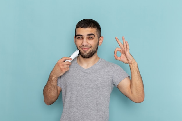 Vue de face jeune homme en t-shirt gris rasant sa barbe sur le bleu glacier