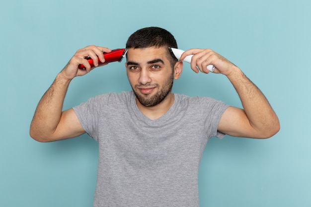 Vue de face jeune homme en t-shirt gris tenant deux rasoirs électriques différents se raser les cheveux sur bleu glace