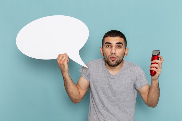 Vue de face jeune homme en t-shirt gris tenant un grand panneau blanc et rasoir électrique sur le bureau bleu glace mâle barbe rasage de cheveux