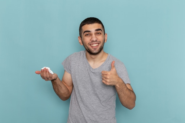 Vue de face jeune homme en t-shirt gris tenant de la mousse blanche pour le rasage souriant sur glace bleu