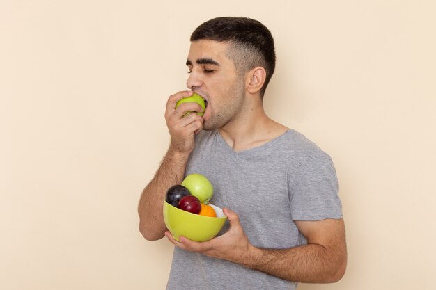 Vue de face jeune homme en t-shirt gris tenant la plaque avec des fruits mordant la pomme sur beige