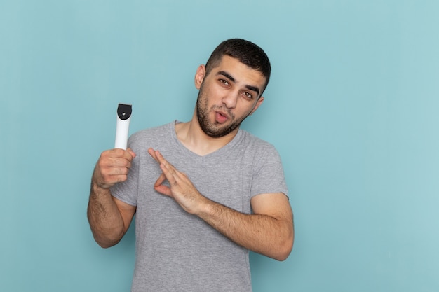 Vue de face jeune homme en t-shirt gris tenant un rasoir électrique sur bureau bleu barbe de rasage mousse de cheveux mâles