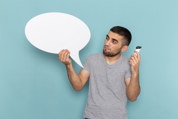 Vue de face jeune homme en t-shirt gris tenant un rasoir électrique avec grand panneau blanc sur homme bleu glace