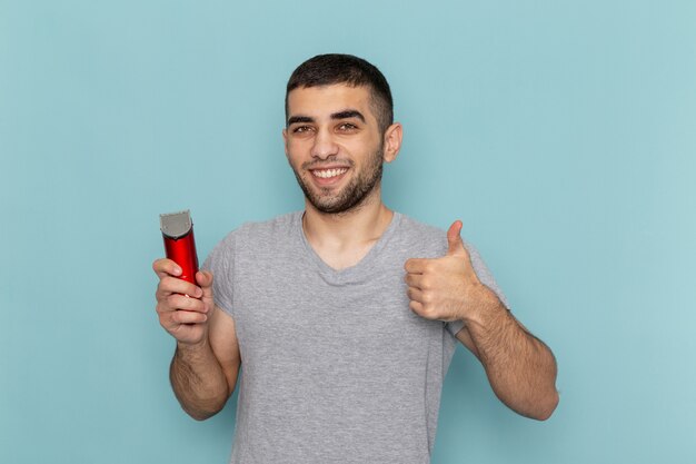 Vue de face jeune homme en t-shirt gris tenant un rasoir électrique montrant un signe impressionnant sur la barbe bleu glace