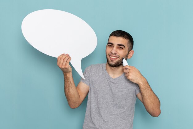 Vue de face jeune homme en t-shirt gris tenant un rasoir électrique avec panneau blanc souriant sur glace bleu