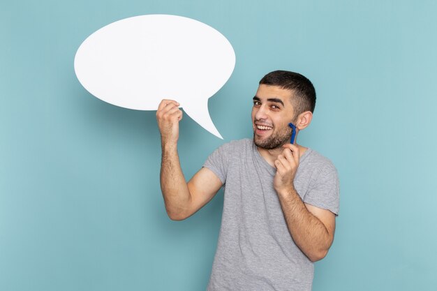 Vue de face jeune homme en t-shirt gris tenant un rasoir et un grand panneau blanc sur le bleu