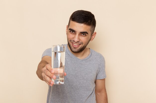 Vue de face jeune homme en t-shirt gris tenant un verre d'eau sur beige