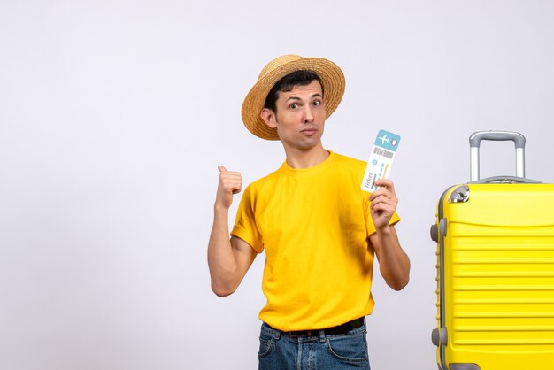 Vue de face jeune homme en t-shirt jaune debout près de valise jaune pointant vers ci-dessous tenant le billet