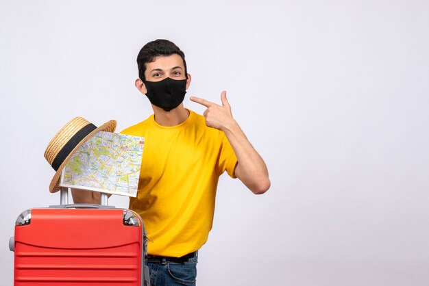 Vue de face jeune homme avec t-shirt jaune et valise rouge pointant sur son masque