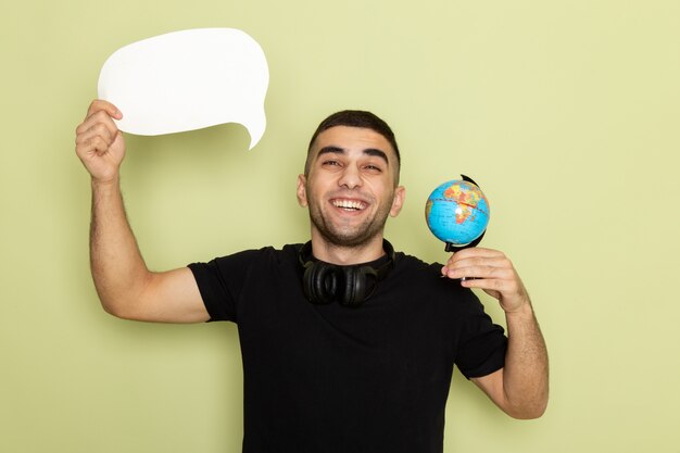 Vue de face jeune homme en t-shirt noir tenant une pancarte blanche et petit globe avec sourire sur vert