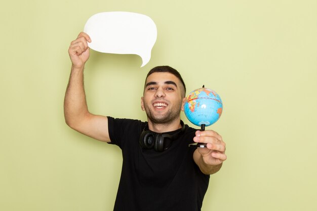 Vue de face jeune homme en t-shirt noir tenant une pancarte blanche et petit globe sur vert