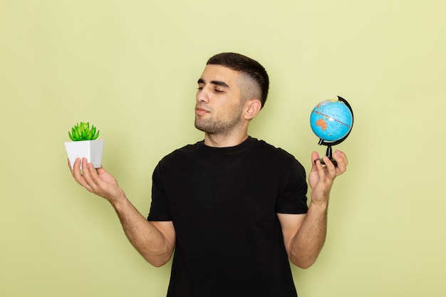 Vue de face jeune homme en t-shirt noir tenant petite plante verte et petit globe sur vert