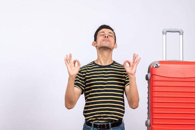 Vue de face jeune homme avec t-shirt rayé et valise rouge faisant signe ok fermant les yeux