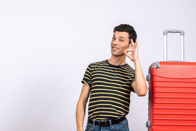 Vue de face jeune homme avec t-shirt rayé et valise rouge faisant signe ok