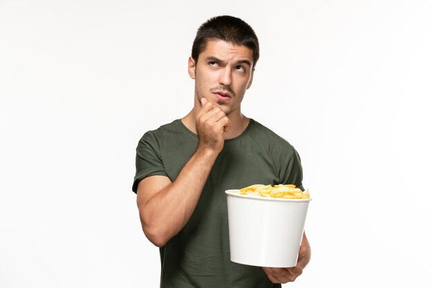 Vue de face jeune homme en t-shirt vert tenant des cips de pommes de terre et regarder un film pensant sur mur blanc cinéma film film solitaire