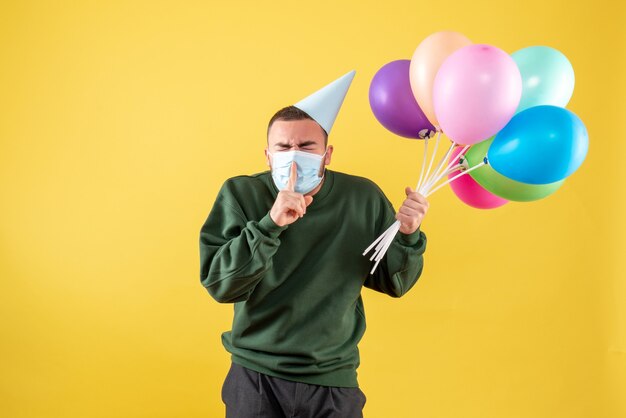 Vue de face jeune homme tenant des ballons colorés en masque stérile sur fond jaune