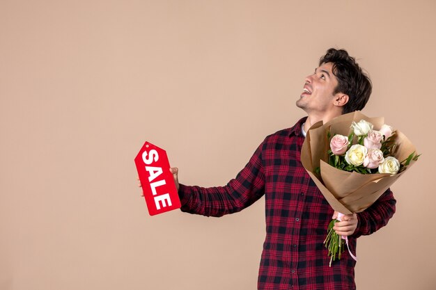 Vue de face jeune homme tenant de belles fleurs et plaque signalétique de vente sur mur marron