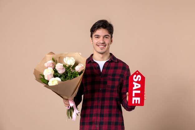 Vue de face jeune homme tenant de belles fleurs pour femme et plaque signalétique de vente sur mur marron