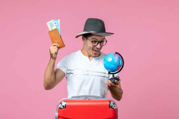 Vue de face d'un jeune homme tenant des billets de vacances et un petit globe sur un mur rose clair