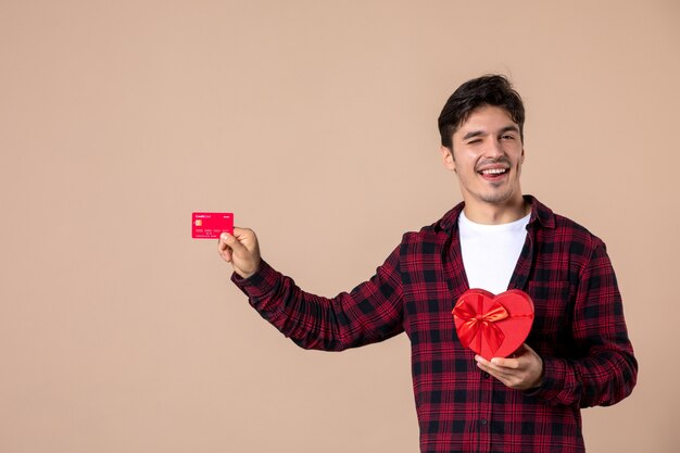 Vue de face jeune homme tenant un cadeau en forme de coeur et une carte bancaire sur un mur marron