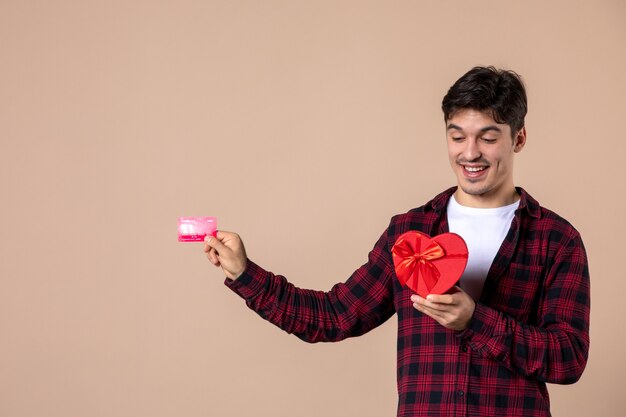 Vue de face jeune homme tenant un cadeau en forme de coeur et une carte bancaire sur un mur marron