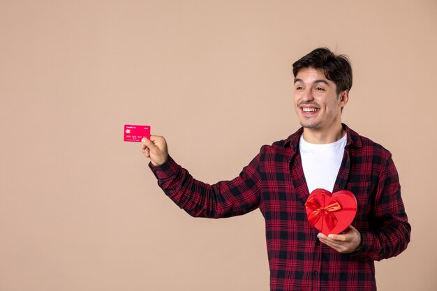 Vue de face jeune homme tenant un cadeau en forme de coeur et une carte bancaire sur un mur marron