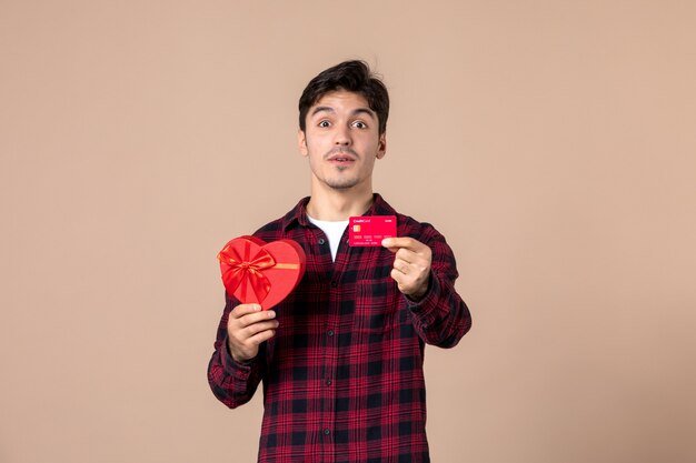 Vue de face jeune homme tenant un cadeau en forme de coeur et une carte bancaire sur un mur marron