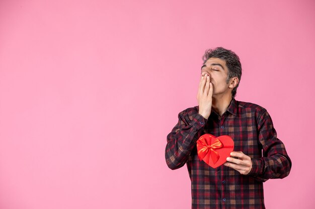 Vue de face jeune homme tenant un cadeau en forme de coeur rouge sur un mur rose