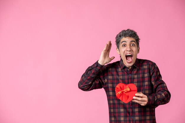 Vue de face jeune homme tenant un cadeau en forme de coeur rouge sur un mur rose