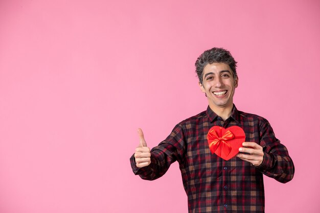 Vue de face jeune homme tenant un cadeau en forme de coeur rouge sur un mur rose