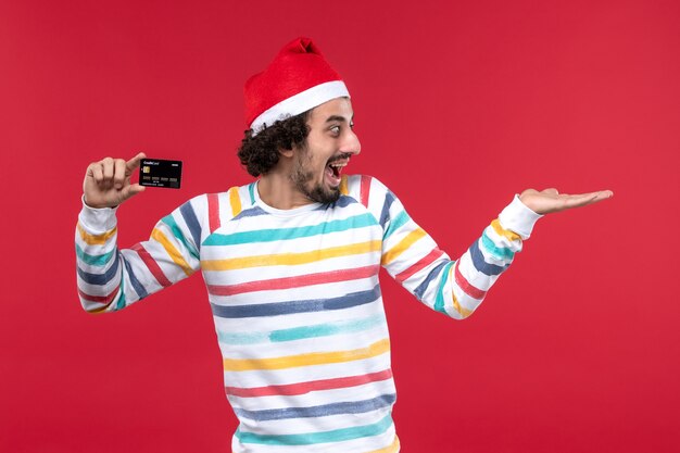 Vue de face jeune homme tenant une carte bancaire sur le mur rouge émotion mâle rouge vacances