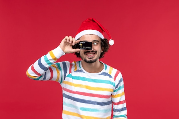 Vue de face jeune homme tenant une carte bancaire sur le mur rouge émotions de vacances mâles rouges