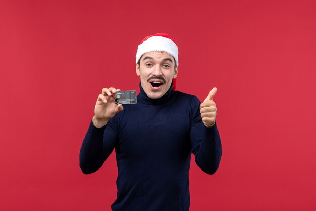 Vue de face jeune homme tenant une carte bancaire noire sur un bureau rouge