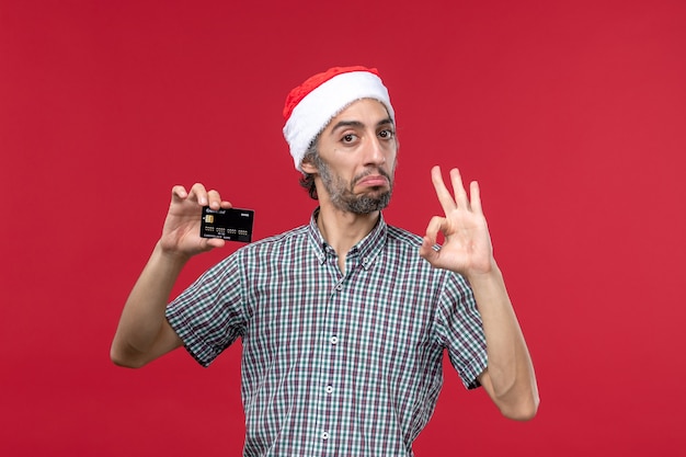 Vue de face jeune homme tenant une carte bancaire noire sur un bureau rouge