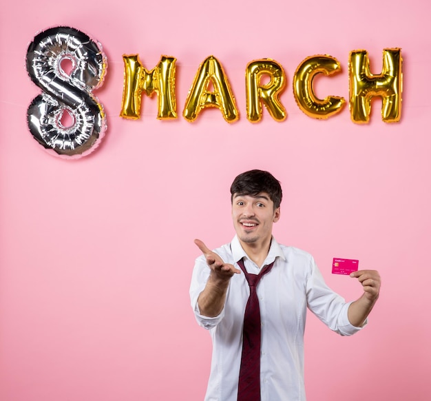 Vue de face jeune homme tenant une carte bancaire rose avec décoration de mars sur fond rose couleur argent fête présente vacances égalité féminine shopping homme