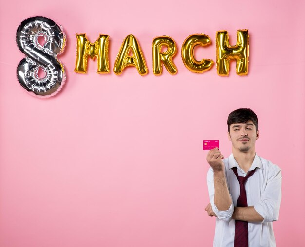 Vue de face jeune homme tenant une carte bancaire rose avec décoration de mars sur fond rose égalité féminine couleur argent shopping homme présent fête
